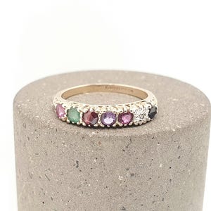 May include: A gold band ring featuring a row of seven round-cut gemstones in various colours including pink, green, red, purple, and black, with a small star-shaped metal detail set between the last two stones on the right side of the band.