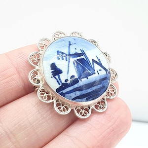May include: A round, vintage brooch with a blue and white landscape design. The porcelain center is framed by a delicate, openwork silver-tone metal border. The brooch depicts a stylized scene with buildings and boats.