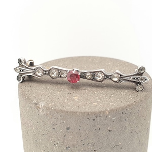 May include: A silver bar brooch featuring a central red gemstone and clear gemstones. The brooch has decorative ends and is displayed on a grey, textured surface. A vintage jewellery piece.