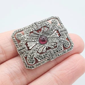 Może przedstawiać: Prostokątna srebrna broszka z centralnym czerwonym kamieniem szlachetnym. Broszka ma bogaty wzór z akcentami markasytu i geometrycznym wzorem. Kamień szlachetny jest okrągły i osadzony we wzorze gwiazdy. Prawdopodobnie vintage lub antyk.