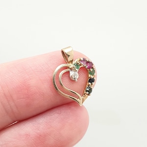 May include: A gold heart-shaped pendant featuring a curved open design. The right side of the heart is accented with a row of five small round gemstones in shades of pink, green, and black, plus a single clear stone set in a silver-toned mount.