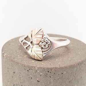 May include: A silver ring with a detailed design featuring leaves and grapes. The leaves have gold and rose gold accents, and the grapes are small and round. The ring is set against a neutral background.
