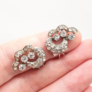 Op de afbeelding: Een paar vintage zilverkleurige clip-oorbellen. Elke oorbel heeft een bloemmotief met heldere, sprankelende strass-steentjes. De oorbellen hebben een klassieke, elegante stijl, perfect om een vleugje glamour toe te voegen.