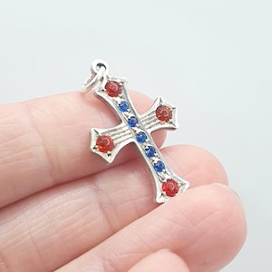 Pode incluir: Pingente de cruz prateada com detalhes de pedras preciosas vermelhas e azuis. A cruz apresenta um design detalhado com uma argola na parte superior para prender a uma corrente. As pedras são dispostas simetricamente.