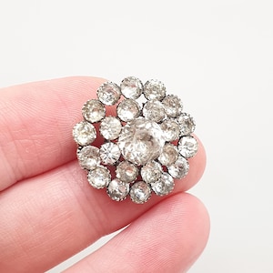 Puede incluir: Un broche circular que presenta una gran piedra central transparente y facetada, rodeada por dos anillos concéntricos de piedras más pequeñas, también transparentes y facetadas, engastadas en un marco de metal oscuro.