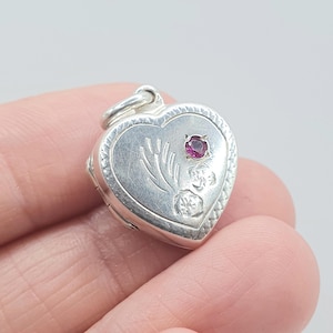 May include: A silver heart-shaped locket with a small, round, red gemstone. The locket has floral engravings and a decorative border. The locket is held in a hand.