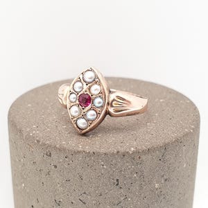 Op de afbeelding: Een marquisevormige ring van roze-getint metaal, voorzien van een centrale ronde rode edelsteen omringd door acht kleine witte parels. De ringband heeft aan beide zijden van de zetting gegraveerde waaiervormige details.