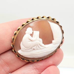 Può includere: Spilla cammeo ovale con cornice color oro. Il cammeo presenta un'incisione a rilievo bianca di una figura seduta e che legge, su sfondo marrone chiaro. La cornice ha un bordo decorativo smerlato.