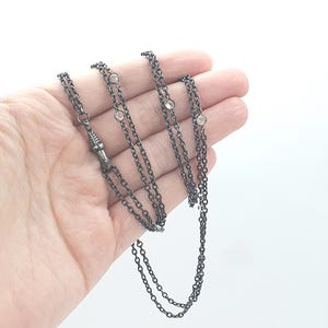 May include: A dark gunmetal chain necklace featuring a series of small, clear, round-cut stones set at intervals along the links. The necklace includes a textured, cylindrical metal clasp mechanism that connects the chain segments together.