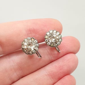 May include: A pair of silver-toned screw-back earrings featuring a central round clear stone surrounded by a halo of smaller clear stones. The circular cluster design is set in a metallic frame with a visible screw mechanism on the back of each piece.