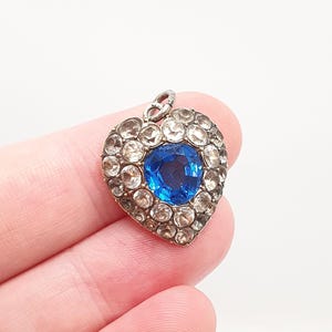 Può includere: Un ciondolo a forma di cuore con una grande pietra centrale blu sfaccettata, circondata da un bordo di pietre più piccole, rotonde e trasparenti, incastonate in una montatura metallica con un piccolo occhiello sulla parte superiore.