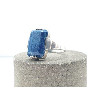 May include: A silver ring featuring a large, rectangular, faceted blue stone with a mottled texture. The metal band has a stepped, geometric design on the sides, leading to a smooth, polished finish that secures the stone in a prong setting.