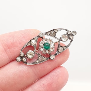 Puede incluir: Un broche alargado de tono plateado con un marco de metal calado engastado con múltiples pedrería redondas transparentes. Un grupo central consiste en una piedra verde esmeralda rodeada por un halo de piedras transparentes más pequeñas.
