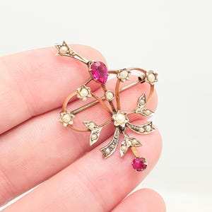 May include: A vintage brooch featuring a floral design with a central teardrop-shaped ruby and smaller round rubies. The brooch is accented with small pearls and has a gold and silver-tone metal finish.