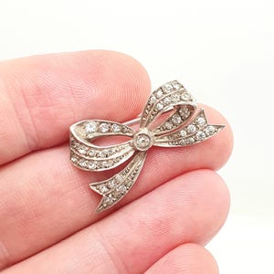 May include: A silver-toned metal brooch shaped like a bow, featuring a central circular stone and multiple small, clear, faceted stones embedded along the ribbon-like structure, creating a sparkling, textured appearance on the metallic surface.