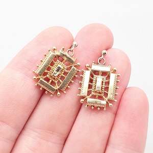 May include: Two gold-toned metal pendants featuring a rectangular central bar surrounded by a geometric frame with textured rope-like detailing and small spherical accents at each corner and along the edges, each with a small circular bail.