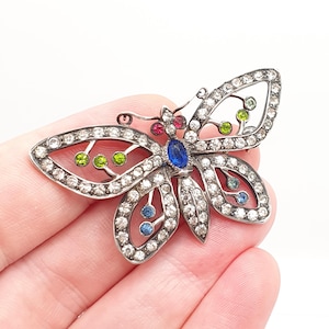 May include: A silver-toned butterfly brooch with clear, blue, green, and red gemstones. The brooch features a detailed openwork design with a central blue gemstone and smaller colored gems throughout the wings.