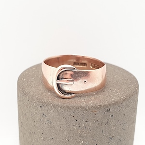 May include: A rose gold ring with a buckle design. The ring is set on a gray surface. The buckle has a curved shape with a small pin. The ring has a polished finish and a vintage aesthetic.