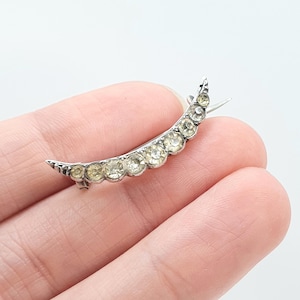 Pode incluir: Um broche em forma de meia-lua em tom prateado com uma fileira de oito pequenas pedras redondas e transparentes facetadas cravadas ao longo da estrutura de metal curva. A peça tem uma ponta em cada extremidade e um mecanismo de alfinete.