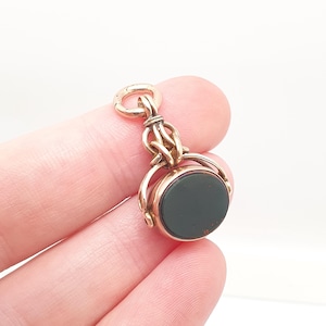 May include: A gold-toned metal swivel fob pendant featuring a circular, dark green stone inlay. The pendant has an ornate, woven metal bail at the top and a rotating mechanism that allows the central stone disc to spin within its frame.