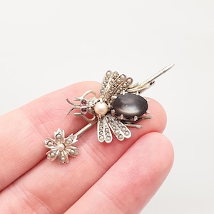 Può includere: Una spilla in argento a forma di insetto con una grande pietra ovale grigio scuro, una perla centrale color crema e piccoli occhi rossi. Le ali e un elemento a forma di fiore sono decorati con minuscole perle. La spilla ha uno stelo lungo e appuntito.