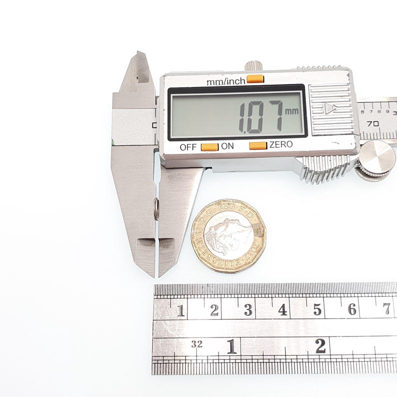 May include: A digital caliper showing a measurement of 1.07 mm, next to a coin and a ruler. The caliper is silver, with a digital display. The ruler is marked with inch measurements. The coin is a British pound coin.