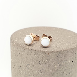 May include: A pair of stud earrings featuring round, smooth white stones set in polished rose gold metal settings. The earrings have standard post backs and are designed with a simple, circular silhouette that highlights the contrast between the white stone and the warm rose gold tone.
