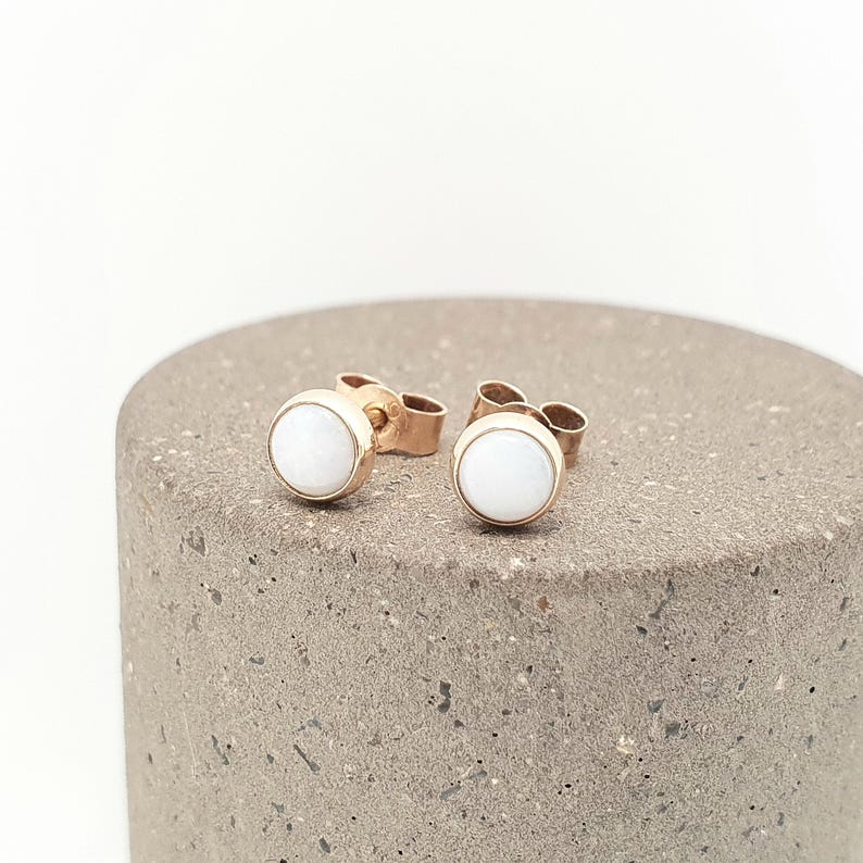 May include: A pair of stud earrings featuring round, smooth white stones set in polished rose gold metal settings. The earrings have standard post backs and are designed with a simple, circular silhouette that highlights the contrast between the white stone and the warm rose gold tone.