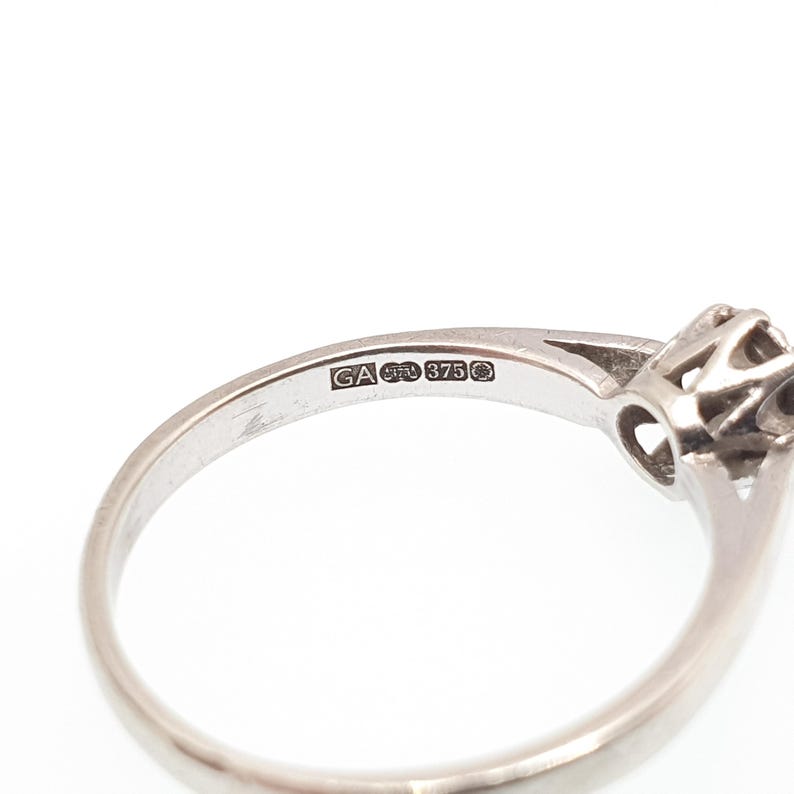Puede incluir: Primer plano de un anillo de plata con una peque&ntilde;a gema redonda. El anillo tiene una banda delicada y un engaste detallado alrededor de la piedra. La marca de contraste "GA 375" es visible en la banda.