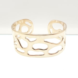 May include: A gold-colored cuff bracelet with an openwork design. The bracelet features irregular, organic-shaped cutouts across its surface. The metal has a polished finish, reflecting light. The bracelet is designed to be worn around the wrist.
