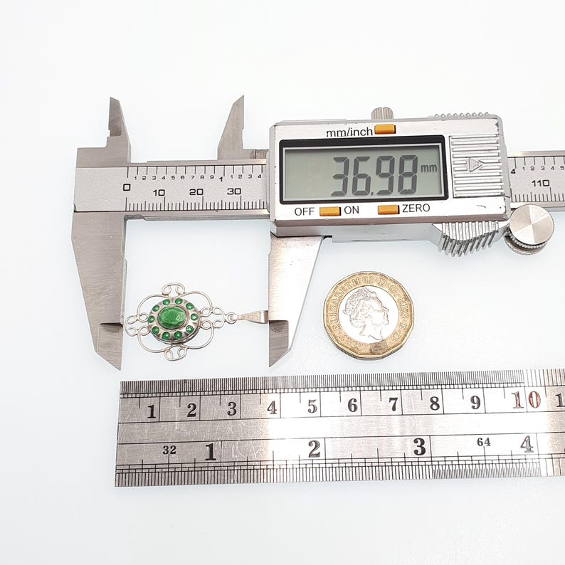 May include: A silver pendant with a central green stone, surrounded by smaller green stones, is measured with a digital caliper. The pendant's length is approximately 1.45 inches. A coin and ruler are also in the image.