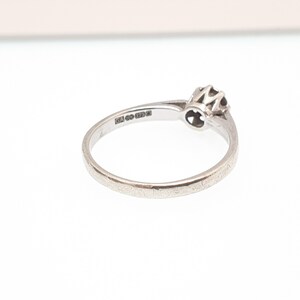 Puede incluir: Un anillo de plata con una peque&ntilde;a gema oscura redonda engastada en un engaste calado decorativo. La banda del anillo es delgada y lisa, con marcas visibles en el interior. El anillo est&aacute; sobre un fondo blanco.