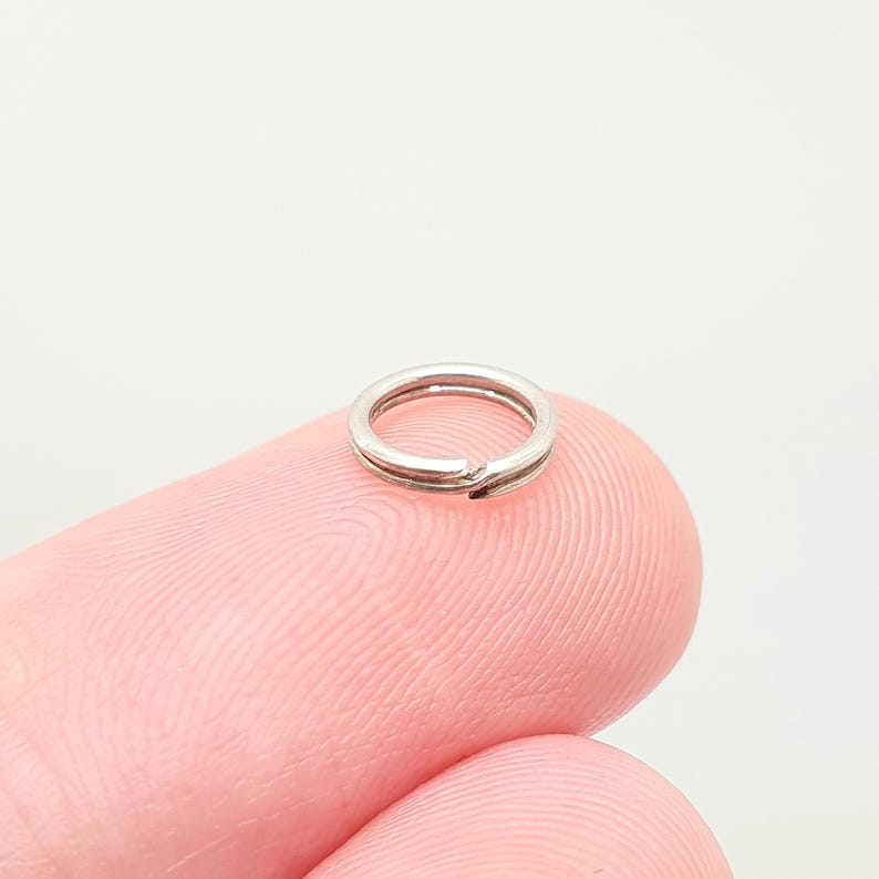 May include: A small, silver split ring, used for attaching items such as keys or charms. The ring is made of metal and has a split design for easy attachment. The ring is shown on a pink finger.