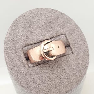 May include: A copper-colored ring with a belt buckle design. The ring features a rectangular band with a small hole and a curved buckle clasp. The ring is displayed on a gray, textured surface.