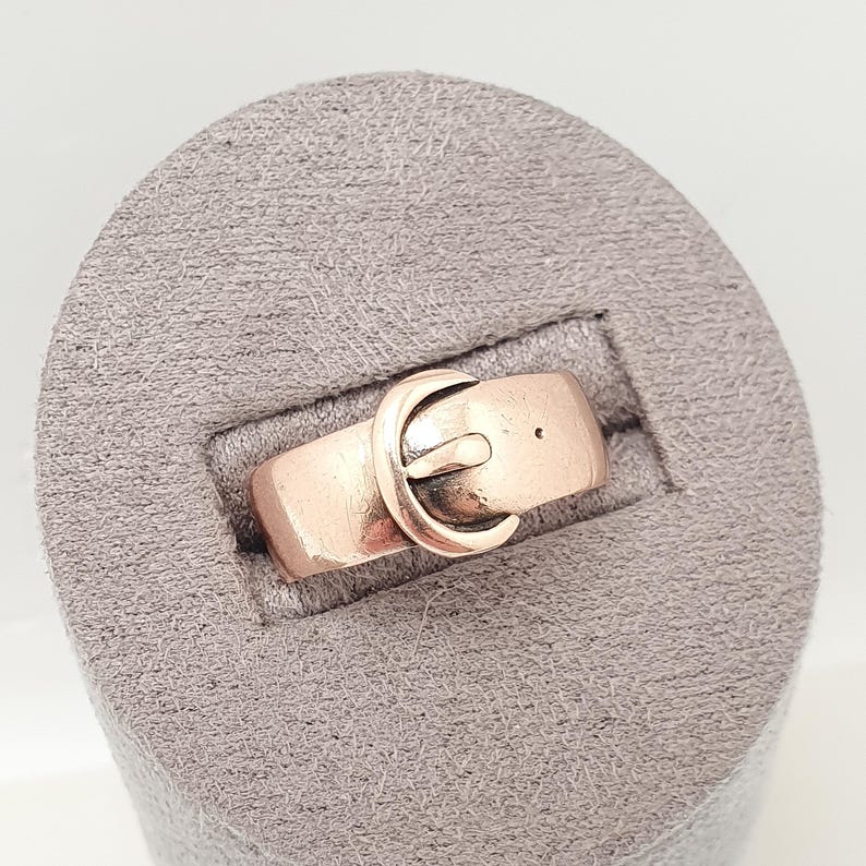May include: A copper-colored ring with a belt buckle design. The ring features a rectangular band with a small hole and a curved buckle clasp. The ring is displayed on a gray, textured surface.