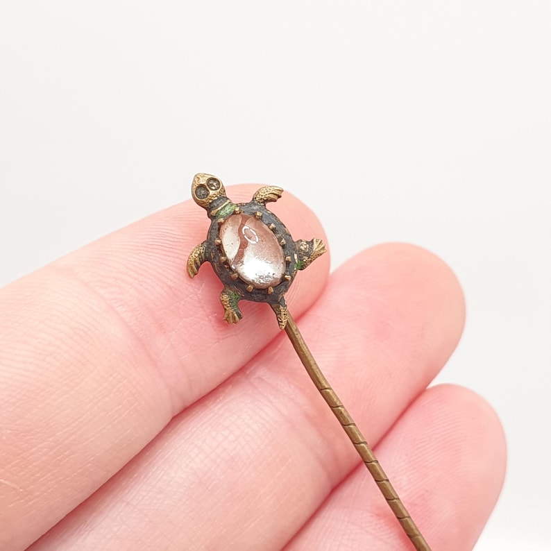 May include: A vintage turtle-shaped pin with a clear, oval-shaped stone set in a bronze-colored metal. The turtle has detailed legs, head, and shell. The pin's stem is a thin, textured bronze color. The pin is likely a piece of jewelry.