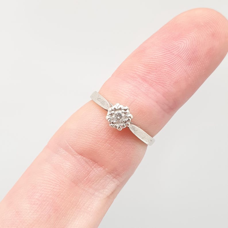 Puede incluir: Un anillo de plata con un engaste en forma de flor y una piedra preciosa transparente en el centro. El anillo se muestra en un dedo, mostrando su delicado dise&ntilde;o y brillo. La banda es fina y elegante.