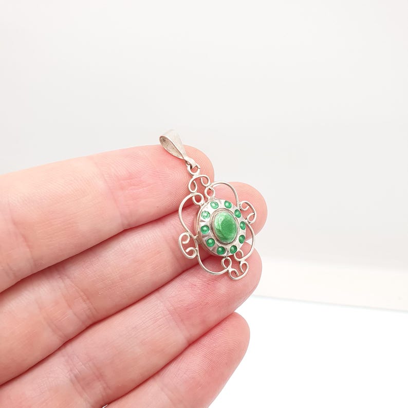 May include: A silver pendant with a floral design. The pendant features a central green stone surrounded by smaller green stones. The pendant has a delicate, openwork design with a loop at the top for a chain.