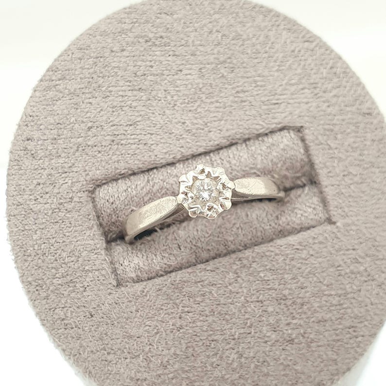 Puede incluir: Un anillo de plata con un diamante redondo central engastado en un dise&ntilde;o floral. El anillo se muestra sobre una superficie gris texturizada. La banda es simple y lisa, lo que conduce al engaste detallado de la piedra preciosa.