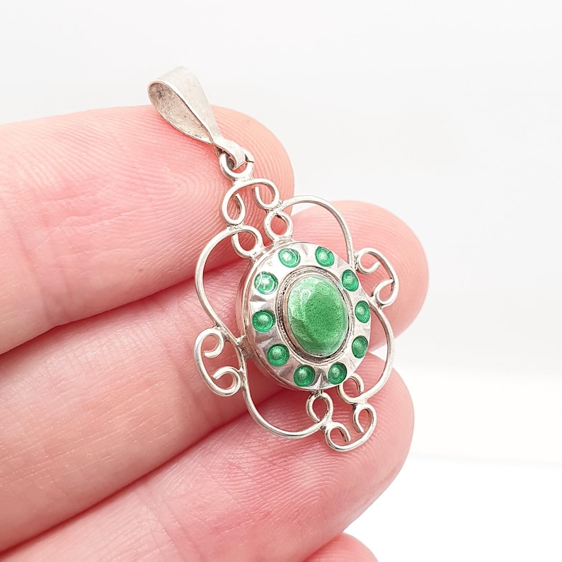 May include: A silver pendant with a floral design. The pendant features a central oval of green enamel surrounded by small green enamel dots. The pendant has a decorative, openwork design with a loop for a chain.