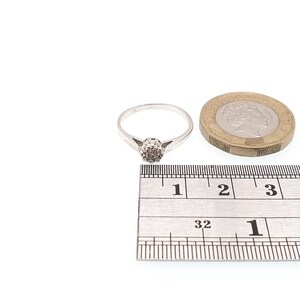 Puede incluir: Un anillo de plata con una peque&ntilde;a piedra redonda de color marr&oacute;n claro en el centro. El anillo est&aacute; junto a una moneda y una regla. La banda del anillo es delgada y se estrecha hacia la piedra.