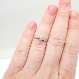 Puede incluir: Un anillo de plata con un grupo de peque&ntilde;as piedras transparentes dispuestas en un patr&oacute;n circular. El anillo se muestra en un dedo sobre un fondo blanco. La banda es delgada y sencilla.