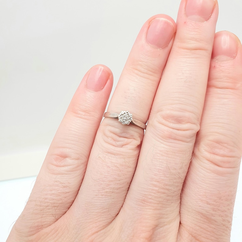 Puede incluir: Un anillo de plata con un grupo de peque&ntilde;as piedras transparentes dispuestas en un patr&oacute;n circular. El anillo se muestra en un dedo sobre un fondo blanco. La banda es delgada y sencilla.