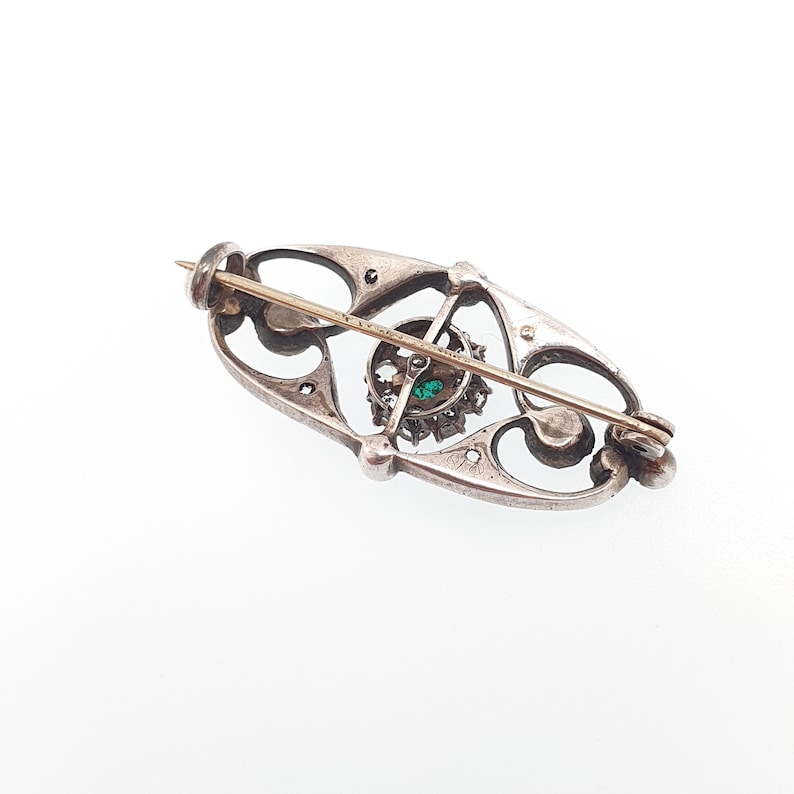 K&ouml;nnte beinhalten: Eine l&auml;ngliche silberne Brosche mit einem durchbrochenen Rahmen und geschwungenen Metalldetails. In der Mitte befindet sich eine kreisf&ouml;rmige Cluster-Fassung mit einem kleinen gr&uuml;nen Stein, umgeben von klaren Steinen und einer horizontalen Nadel.