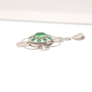 May include: A silver pendant with a circular green stone center and green enamel accents. The pendant has a decorative, openwork design and a loop for a chain. The pendant is likely made of sterling silver.