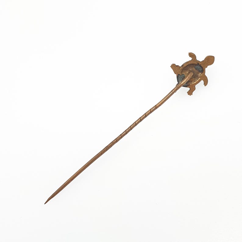 May include: A vintage bronze hat pin featuring a turtle design. The pin has a long, slender shaft with a spiral detail near the top. The turtle is detailed with visible legs, head, and shell. The pin is set against a white background.