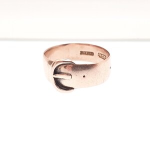 May include: A rose gold ring in the shape of a belt buckle. The ring has a classic buckle design with a small pin and a series of holes. The ring is set against a white background.