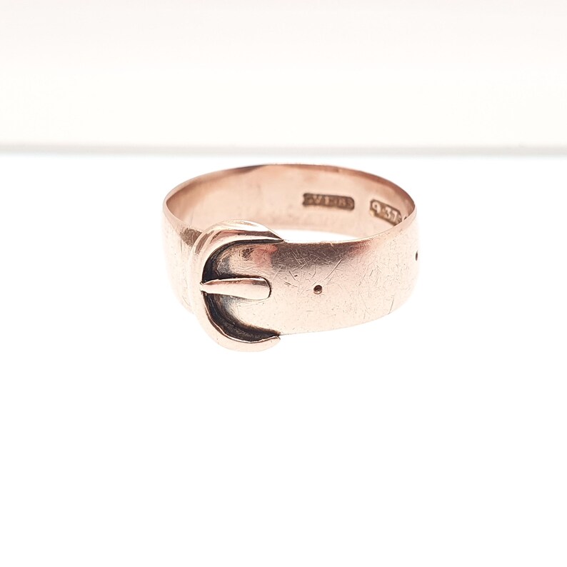 May include: A rose gold ring in the shape of a belt buckle. The ring has a classic buckle design with a small pin and a series of holes. The ring is set against a white background.