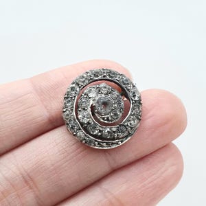 Pode incluir: Um broche circular de tom prateado com um design em espiral incrustado com numerosas pedras pequenas, transparentes e facetadas. A peça é construída com uma pedra central rodeada por uma banda contínua de pedras correspondentes.
