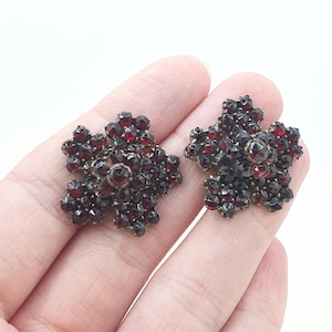 Op de afbeelding: Twee stervormige cluster-oorbellen met meerdere gefacetteerde dieprode granaatstenen in een donker metalen frame. Elke oorbel heeft een verhoogde centrale steen omringd door kleinere stenen in een bloemachtig patroon met metalen achterkant.